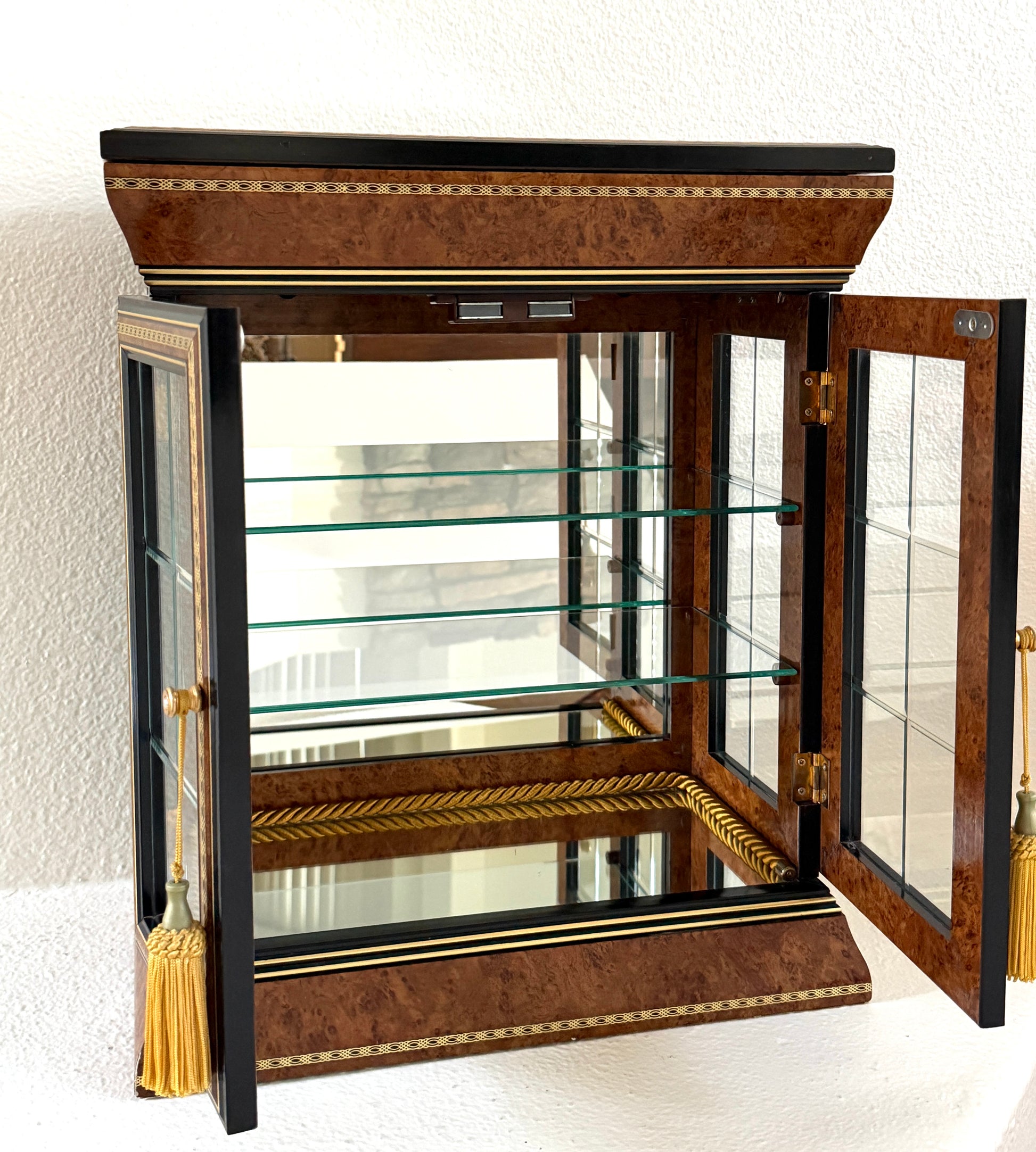 A vintage-style display cabinet with two glass doors, featuring a rich brown burl wood veneer, black trim, gold geometric inlay, and gold tassels on the door handles made in Italy