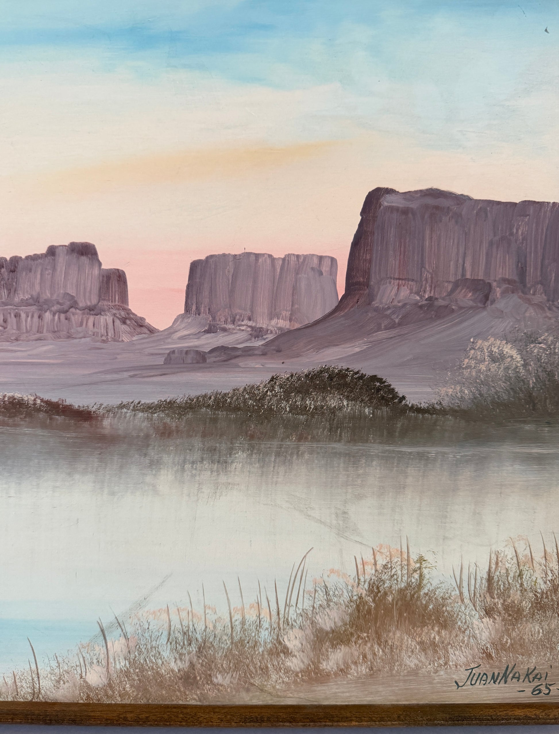A painted landscape scene showing a calm lake or river in the foreground with grasses, misty trees, and large rock buttes or mesas in the background under a pale blue and pink sky. The artwork appears to be signed in the lower right corner.