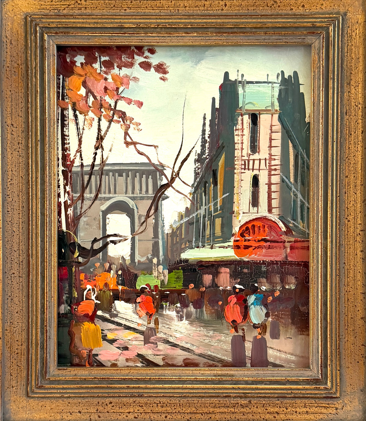 A framed oil painting depicting a busy city street scene with pedestrians, storefronts, trees with autumn leaves, and a bridge in the background, enclosed in a textured gold-toned frame.