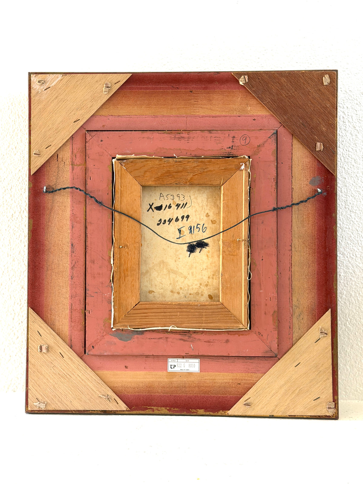 The back of a square wooden picture frame with layered reddish-brown and natural wood sections, a hanging wire, and handwritten identification markings in the center