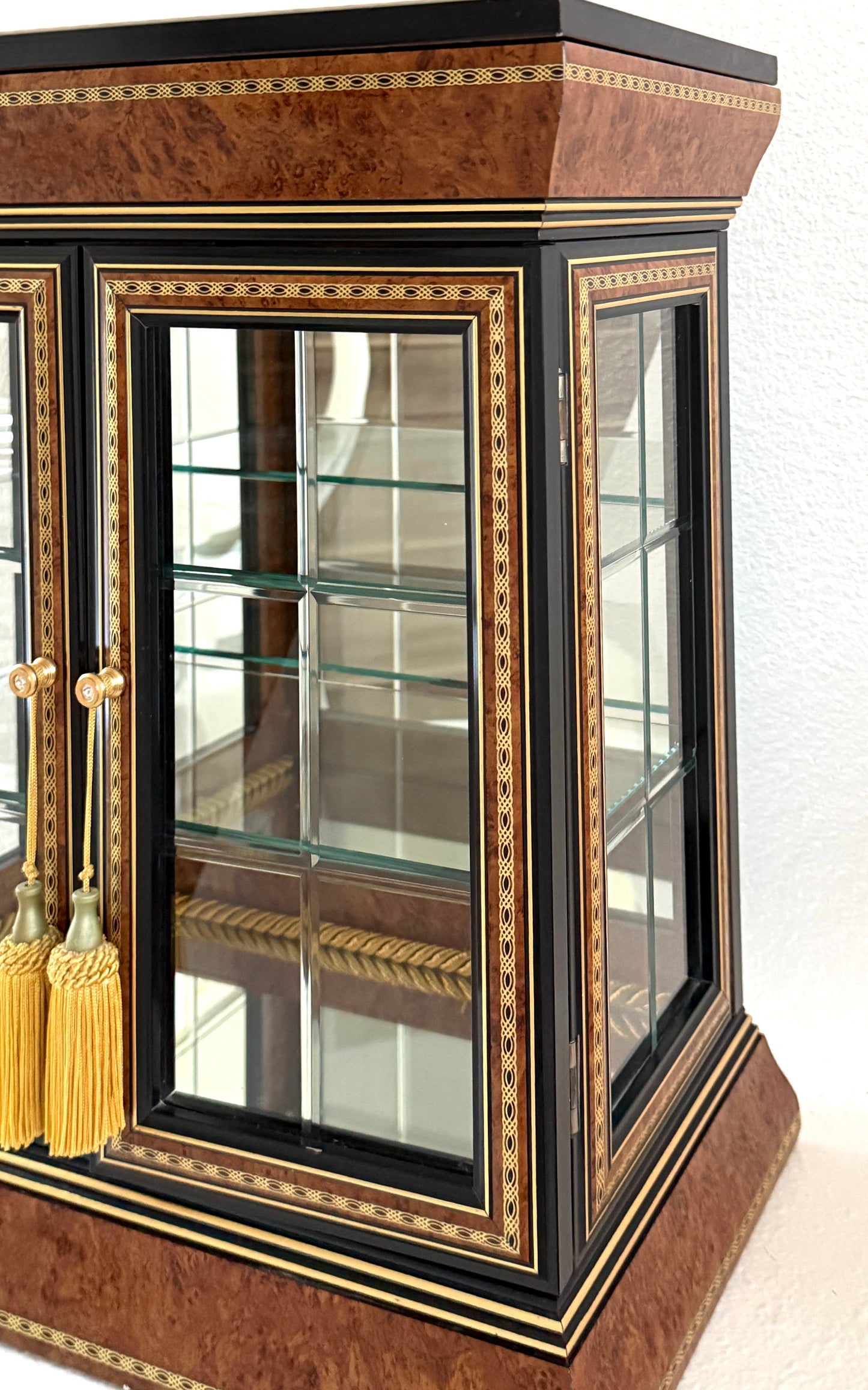 A vintage-style display cabinet with two glass doors, featuring a rich brown burl wood veneer, black trim, gold geometric inlay, and gold tassels on the door handles made in Italy