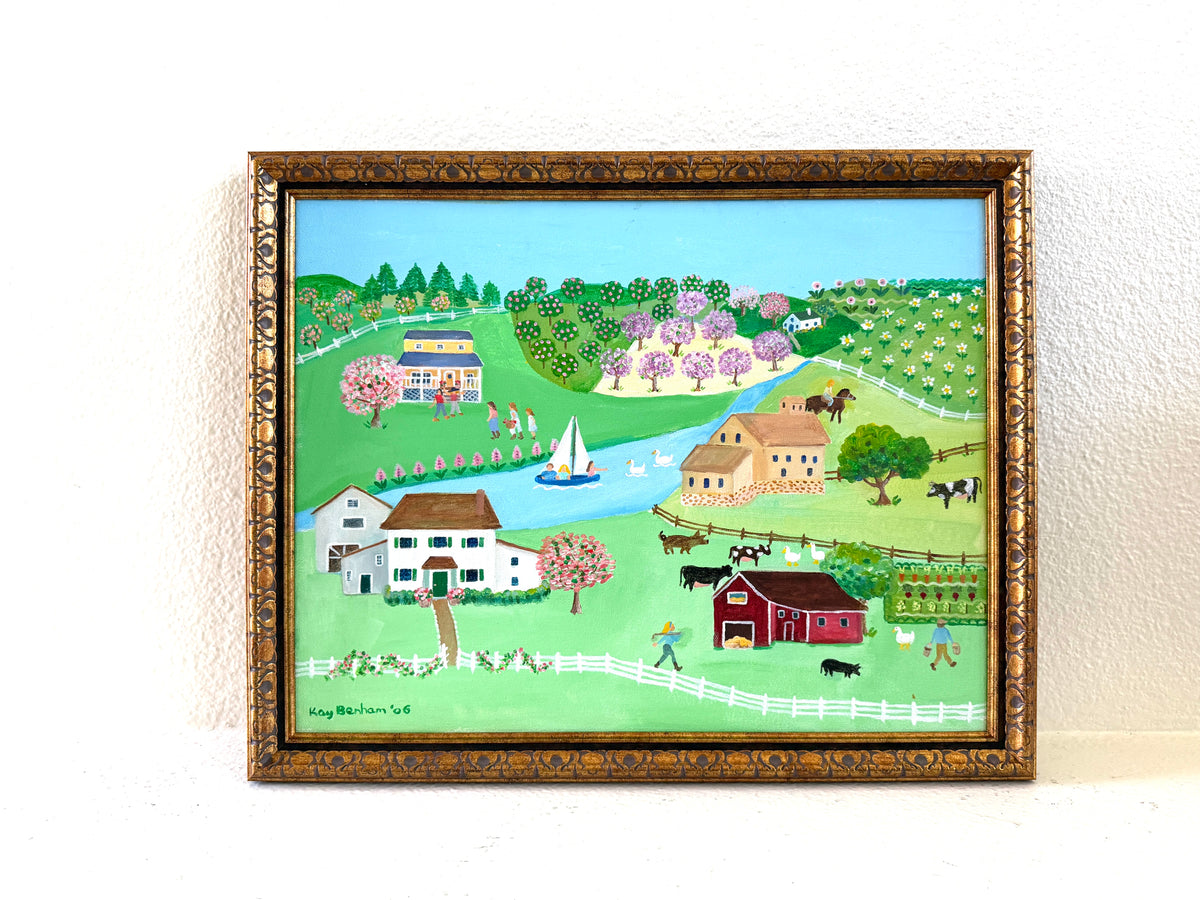 A framed folk art landscape painting depicting a rural scene with houses, barns, trees, fields, and a river Signed Kay Benham 06