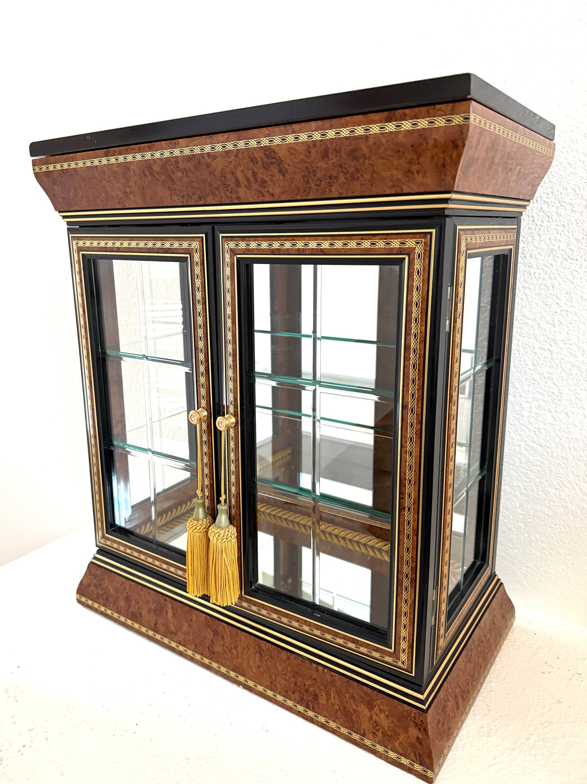 An ornate Art Deco style glass display cabinet with a rich brown burl wood veneer, black lacquered trim, gold geometric inlay, and gold tassels on the doors, Made in Italy 