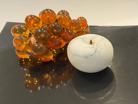 Artificial amber-colored grapes and a white marble apple on a reflective black surface.