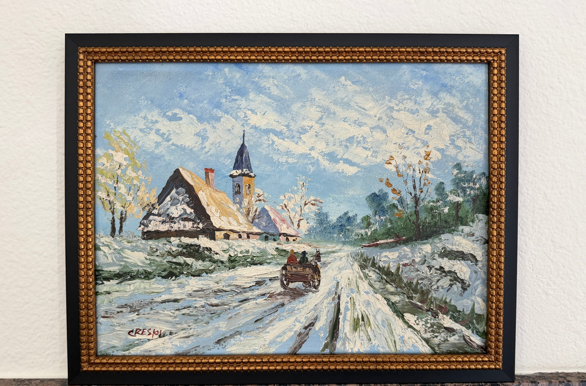 A framed winter landscape painting depicting snow-covered roads, traditional houses, trees, and a church steeple under a cloudy blue sky, set in a decorative gold-toned frame against a white wall.