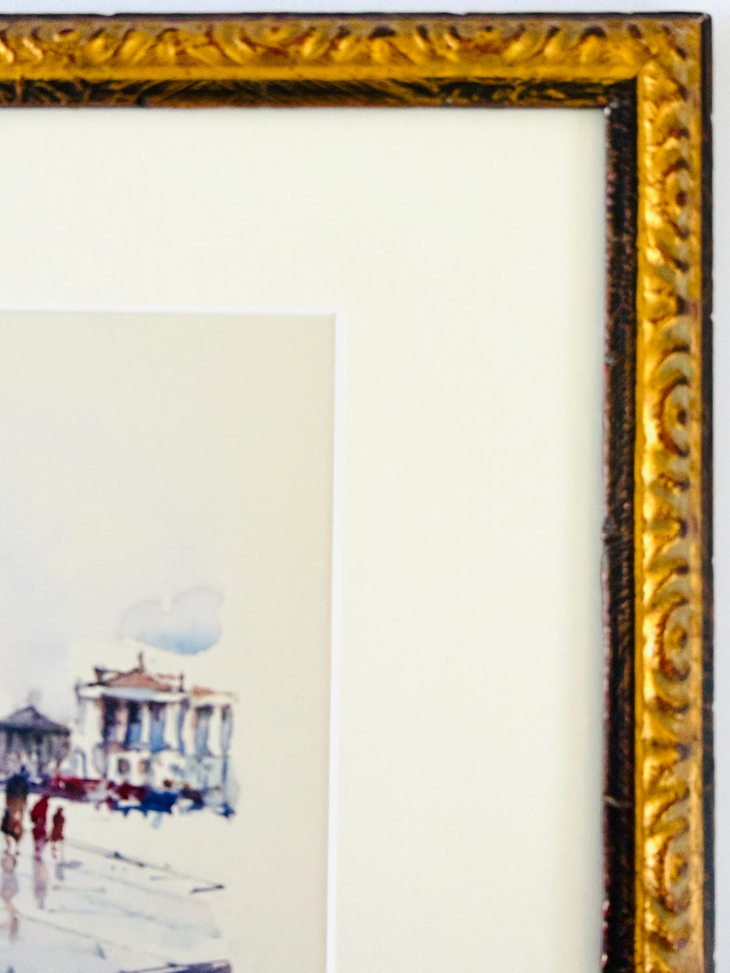 Close-up of an ornate gold picture frame with a white mat and a partial watercolor painting showing a seaside pavilion scene.