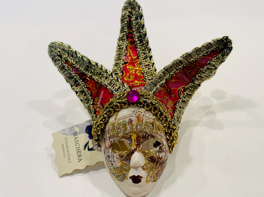 A decorative Venetian mask featuring gold, pink, and red colors, with textiles and ceramic details, placed on a white surface with a card attached.